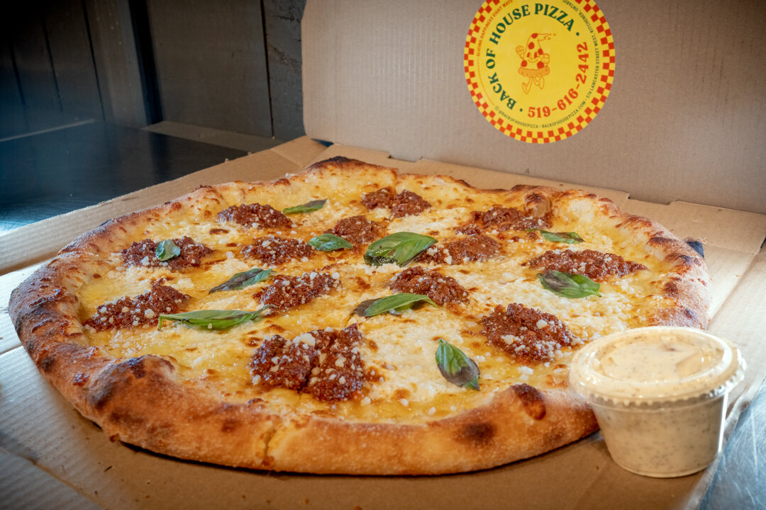  A Back of House Pizza box opened to reveal the Bolognese pizza, topped with rich sauce, melted cheese, and fresh basil, with a house dipping cup beside it and the Back of House Pizza logo visible on the lid.